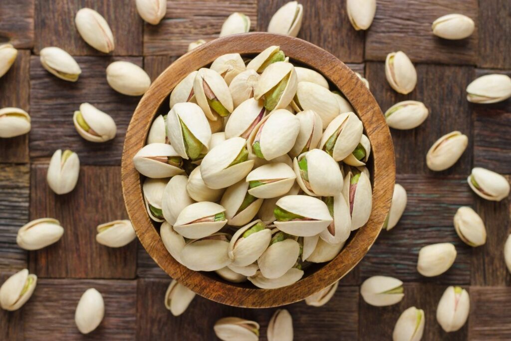 A bowl of pistachios is surrounded by unshelled pistachios